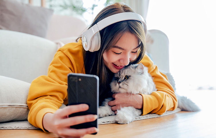A young woman wearing headphones and a yellow sweatshirt lies on the floor, cuddling her fluffy gray cat while taking a selfie. Concept for cat pet insurance.